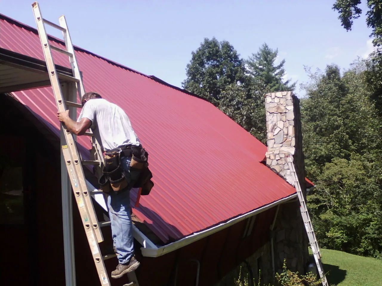 Roofer on a ladder beside a red metal roof and stone chimney — Soffit & Fascia Repair by Roofing Brothers in Bridge City